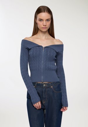 Maglione blu scollato con zip, caratterizzato da una texture a treccia, polsini a coste e un design aderente, abbinato a jeans blu scuro.