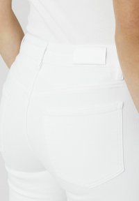 Jeans en denim blanc avec une silhouette ajustée, comportant des poches arrière, des passants de ceinture et une texture lisse. Aucun logo ou motif visible.