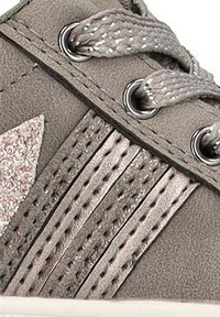Zapatilla gris con un material texturizado similar al ante, acentos metálicos, cordones trenzados y un logo lateral brillante. Presenta ojales plateados y detalles a rayas.