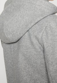 Abrigo gris con capucha hecho de tela suave y texturizada. Presenta una capucha redondeada y detalles de costuras en los hombros. Diseño simple y minimalista.