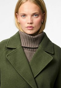 Oliivivärvi villane jope, millel on teravad kraed, kihistatuna pruunist ribakudumist sõrmkuubist, millel on tekstureeritud kudumine ja kõrge kaelus.