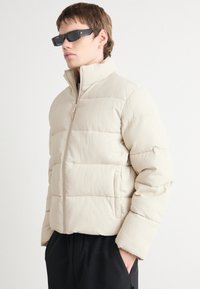 Jachetă puffer bej cu textură ribbed, guler înalt, închidere cu fermoar și manșete elastice; stilizată cu ochelari de soare negri, rectangulari.