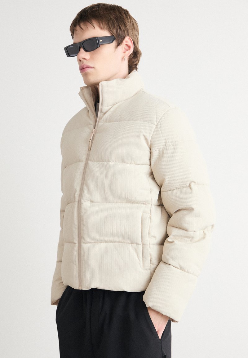 Jachetă puffer bej cu textură ribbed, guler înalt, închidere cu fermoar și manșete elastice; stilizată cu ochelari de soare negri, rectangulari.