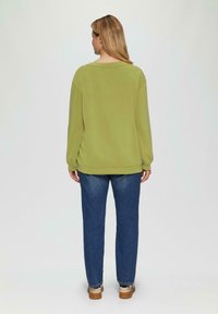 Vrouw staand, gekleed in een lichtgroen shirt met lange mouwen, blauwe jeans en bruine schoenen, met de rug naar de camera gekeerd tegen een effen achtergrond.