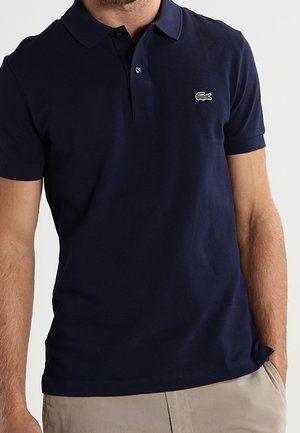 Homme portant un polo à manches courtes bleu marine avec un logo de crocodile vert sur la poitrine gauche et un pantalon beige.