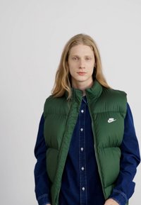 Grøn polstret vest over en blå denimskjorte, med høj krave og et hvidt Nike-logo på venstre brystområde. Glat tekstur.