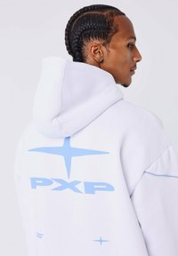 Jonge man met gevlochten haar, gekleed in een witte hoodie met blauwe sterontwerpen en de tekst "PXP", kijkend naar rechts.