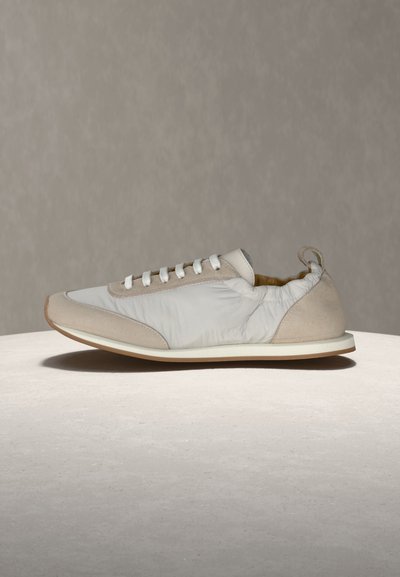 Ανοιχτό γκρι και μπεζ αθλητικό παπούτσι με λευκά κορδόνια, μαξιλαρωμένο γιακά στη φτέρνα και επίπεδη σόλα πάνω σε ελαφρύ στρογγυλό υπόστρωμα με απλό φόντο.