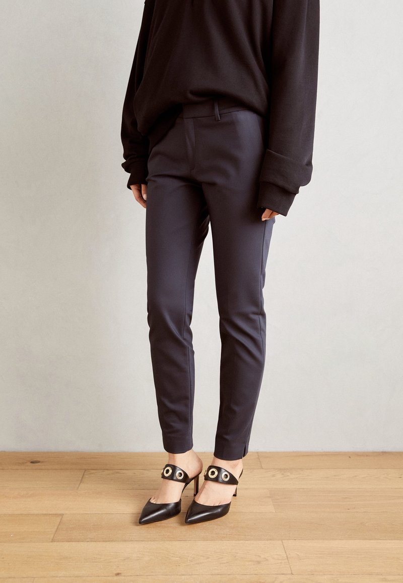 Black oversized sweater, fitted dark navy pants, and black pointed-toe heels with circular accents on the straps, set on a wooden floor.