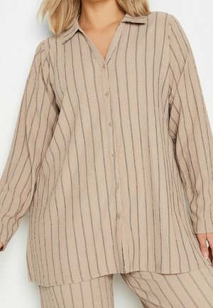 Beżowa koszula na długi rękaw z guzikami, z czarnymi pionowymi paskami, noszona z pasującymi beżowymi spodniami w paski.