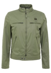 Veste vert olive en matériau lisse et léger. Elle est dotée d'un col montant, de deux poches poitrine zippées et d'un ourlet côtelé.