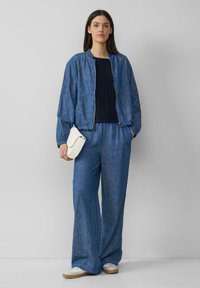 Denim bomber jacket dengan manset elastis, dipadukan dengan celana biru yang longgar. Model memegang clutch putih, mengenakan sneaker beige.