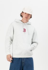 Felpa grigia con tasca frontale, caratterizzata da un logo grafico color bordeaux. Abbinata a jeans a gamba larga blu e un berretto color crema.