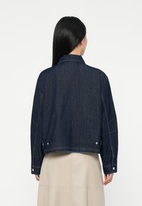 Kvinne med langt svart hår iført en mørkeblå denimjakke og et beige skjørt, vist fra bakgrunnen mot en enkel hvit bakgrunn.