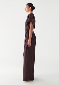 Largo vestido marrón hecho de una tela suave, con una cintura ajustada, mangas cortas y una silueta recta con un lado ligeramente abierto.