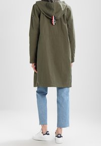Olivově zelená lehká bunda s kapucí, s úzkým pasem a detailem na zadní straně. V kombinaci se světle modrými zkrácenými džíny a bílými teniskami.
