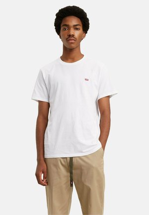 Jonge man met een afro kapsel, gekleed in een wit Levi's T-shirt en beige broek met een groene riem, staand tegen een eentonige achtergrond.