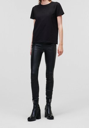 Personne portant un t-shirt noir à manches courtes, un pantalon en cuir noir et des bottines noires à talons avec fermetures éclair à l'avant, debout devant un fond uni.