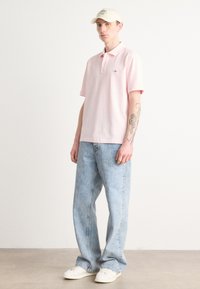 Polo rose clair avec col et manches courtes, associé à un jean en denim bleu, à coupe ample, et des baskets blanches. Le chapeau est beige avec du texte.