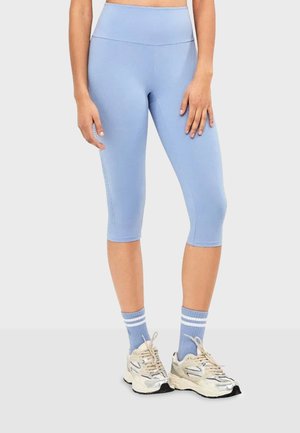 Persona que lleva mallas cortas azul claro, calcetines azules a juego con rayas blancas y zapatos deportivos beige, de pie sobre un suelo gris.