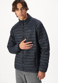 Czarna kurtka puchowa z pikowanym wzorem, wysokim kołnierzem, zamkiem błyskawicznym z przodu oraz bocznymi kieszeniami. Posiada subtelną łatkę na lewej piersi.