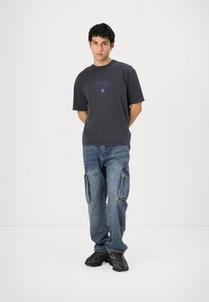 Grå bomull T-skjorte med "GUESS" logo i blått over et trekantdesign, sammen med falmede blå cargo jeans med flere lommer.
