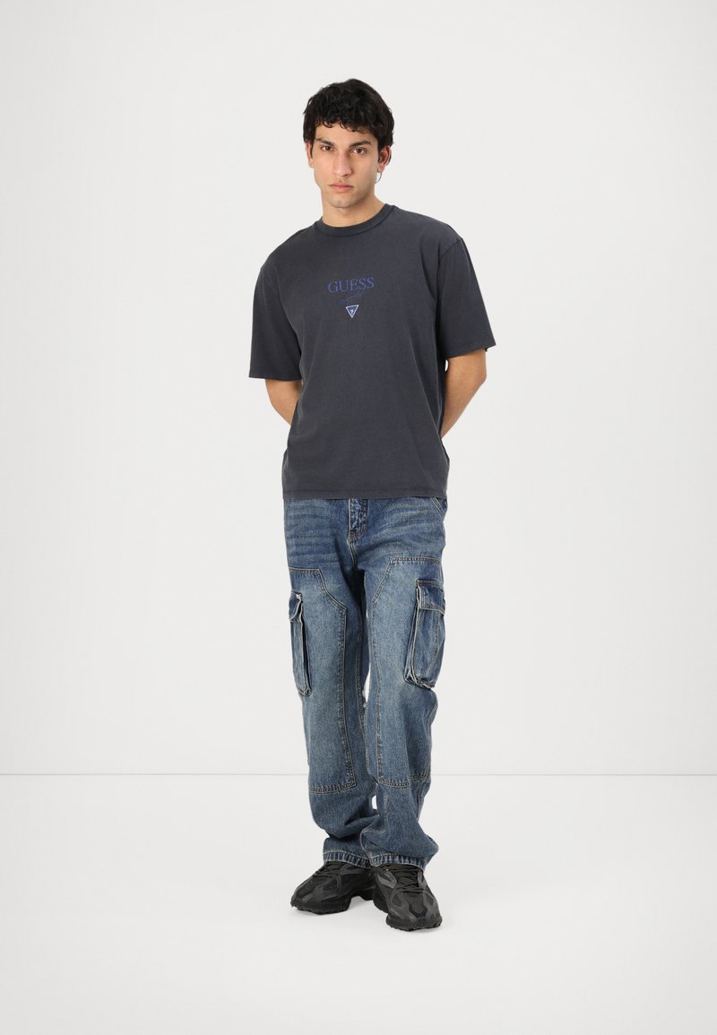 Camiseta de algodón gris con el logotipo "GUESS" en azul sobre un diseño de triángulo, combinada con jeans cargo azules descoloridos que cuentan con múltiples bolsillos.