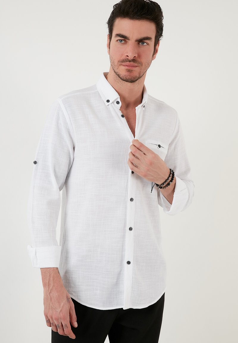 Camicia bianca a maniche lunghe in tessuto texture, con bottoni neri e taschino sul petto. Dettagli del polsino e del colletto abbottonati visibili.
