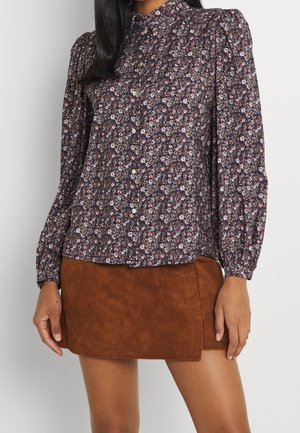 Femme portant un chemisier floral à manches longues boutonné avec des épaules bouffantes et une mini-jupe en suède marron.