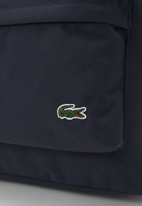 Bolsa de tela azul marino con una superficie texturizada y un pequeño logo bordado de cocodrilo verde con un acento rojo.