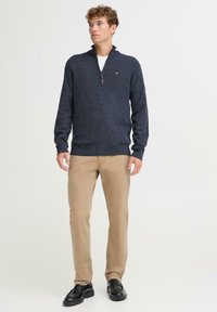 Suéter de punto azul marino con cuello con cremallera, puños acanalados y dobladillo. Combinado con pantalones caqui y zapatos de vestir negros. Textura de tela suave.