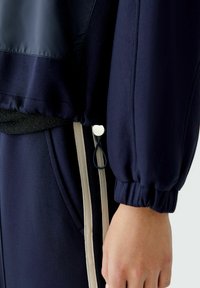 Marine trainingsjack met een glanzend bovenpaneel, elastische manchetten en een verstelbaar trekkoord aan de onderkant, met crèmekleurige zijstrepen.