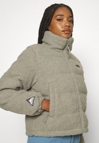 Giacca in pile con zip di colore grigio chiaro, colletto alto, tasche laterali e patch del logo sulla manica sinistra. Materiale strutturato e caldo.