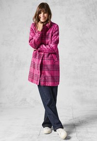 Fuchsia ternet frakke med krave, knaplukning foran og sidelommer. Blødt, struktureret stof sammen med mørkeblå jeans og hvide sneakers.