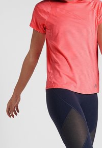 Krátký rukáv sportovní top v jasně korálové barvě s horizontálními pruhy, vyrobený z lehkého materiálu, spárovaný s tmavými legínami s síťovinovými akcenty.