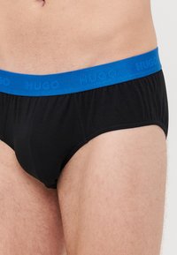 Zwarte boxershorts met een blauwe elastische tailleband waarop de merknaam "HUGO" is verdiept. De stof lijkt glad en rekbaar.