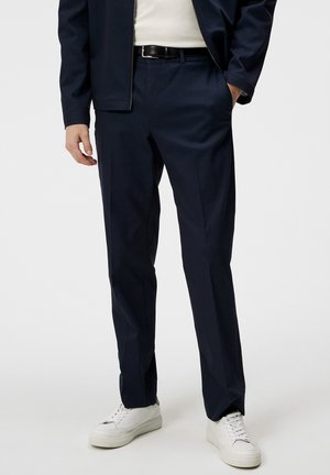 Marineblauwe broek met een getailleerde pasvorm, gemaakt van zachte stof; voorzien van zakken en een subtiele plooi aan de voorkant. Gecombineerd met witte sneakers.