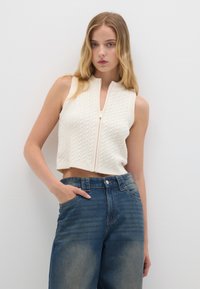 Gilet lavorato a maglia color crema con motivo a treccia, lunghezza corta e zip frontale, abbinato a jeans in denim blu a vita alta.