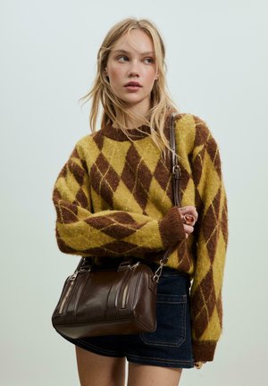 Jeune femme portant un pull en argyle marron et jaune, une jupe en denim, tenant un sac à main en cuir marron foncé avec une bandoulière.