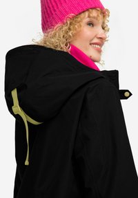 Veste noire avec capuche, dotée d'une sangle ajustable verte fluo. Superposée avec un col rose vif, fabriqué en tissu doux avec une texture lisse.
