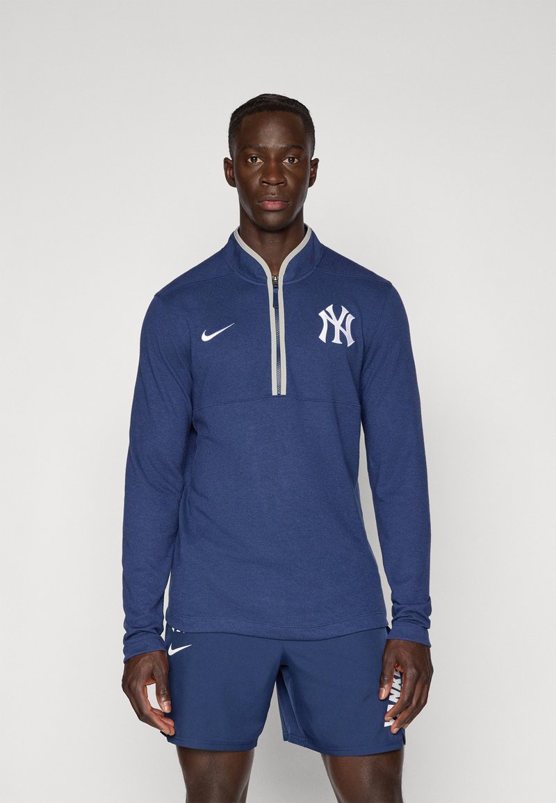 Námořnická modrá košile s dlouhým rukávem a zipem, s šedým límcem, na hrudi s výrazným bílým logem NY a swoosh Nike. Vyrobena z texturované látky.