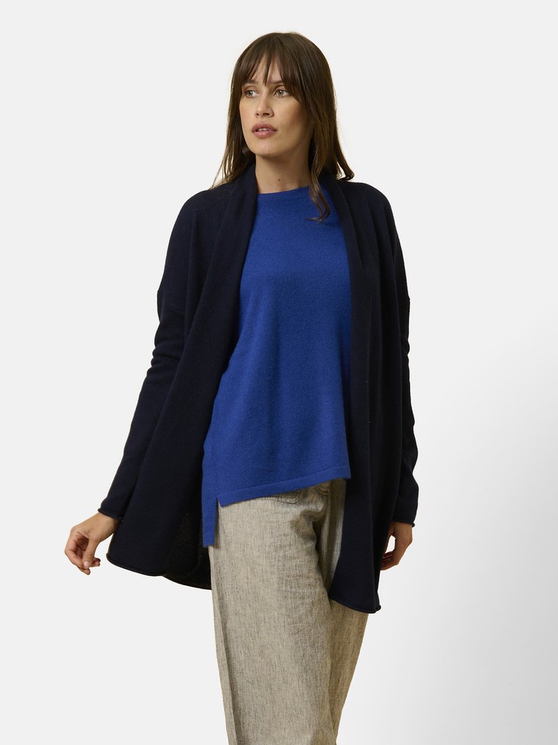 Kvinna klädd i en marinblå öppen cardigan, blå tröja med rund hals och beiga vida byxor, stående mot en enkel ljus bakgrund.