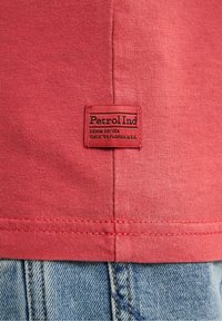 Červené bavlněné tričko s černou nášivkou "Petrol Ind", viditelné švy a detaily lemu. Nosené s denimovými džíny.