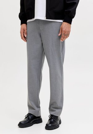 Homme portant un pantalon gris clair, des chaussures noires en cuir à lacets, une veste noire et une chemise blanche, debout sur un fond clair uni.