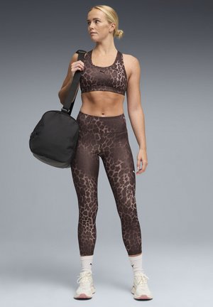 Top deportivo con estampado de leopardo marrón y leggings a juego, combinados con una bolsa de gimnasio negra. La modelo lleva zapatillas deportivas claras y calcetines blancos hasta la mitad de la pantorrilla.