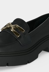 Mocassin en cuir noir avec semelle épaisse et détail de boucle en métal doré sur le dessus, vue rapprochée de l’avant et du côté.