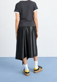 Osoba stojící na světle modré podlaze, oblečená v černém tričku s krátkým rukávem, asymetrické černé sukni, bílých ponožkách a černých a žlutých teniskách.