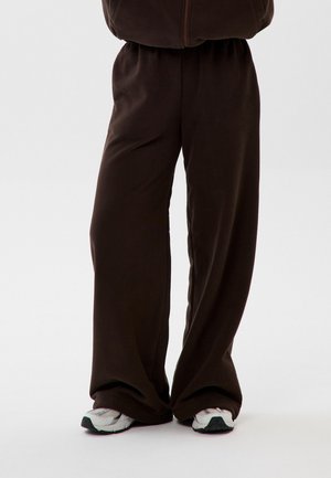 Osoba nosząca luźne ciemnobrązowe spodnie i białe trampki, stojąca na jednolitym białym tle.