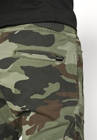 Vihreän sävyiset maastokuvioiset cargo-housut, joissa on ruskeat yksityiskohdat, sivutasku ja musta "Hollister"-etiketti takana.