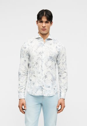 Άνδρας που φοράει λευκό πουκάμισο με μακρύ μανίκι και κουμπιά, με ανοιχτό γκρι σχέδιο παϊλετί και ανοιχτούς μπλε τζιν, στέκεται μπροστά σε απλό φόντο.